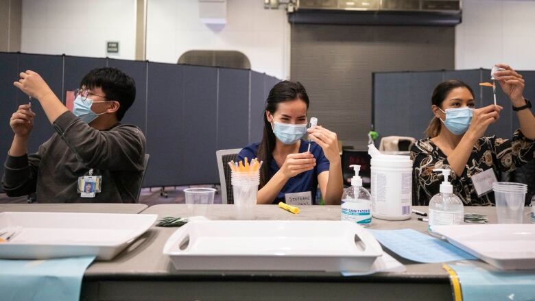 Three workers in medical masks load up syringes with vaccines.