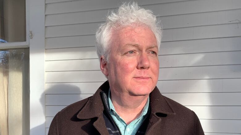 A man with white hair wearing a brown jacket stands in front of a barn.