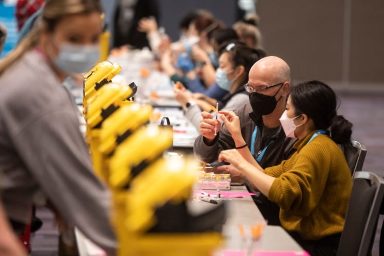 People wearing masks sit at a long table, hold syringes.