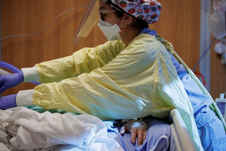 A woman in full PPE leans over a patient on a ventilator.