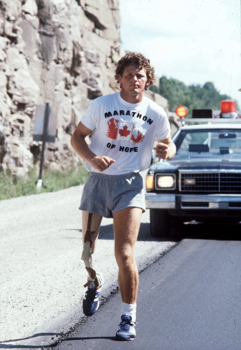 Terry Fox runs along a road with a car behind him. He is wearing a shirt that reads 'Marathon of Hope' and has a prosthetic leg.