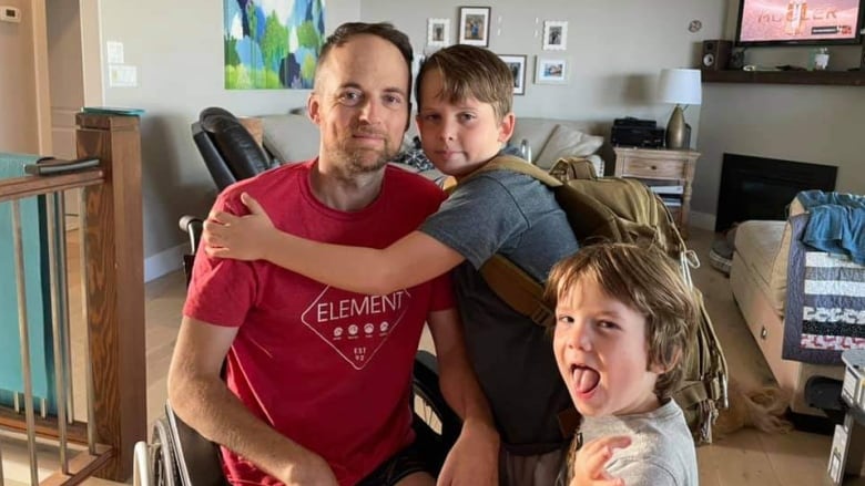 A man in a maroon T-shirt in a room at home with a child on his lap hugging him looks at the camera while one of his other children makes a face.