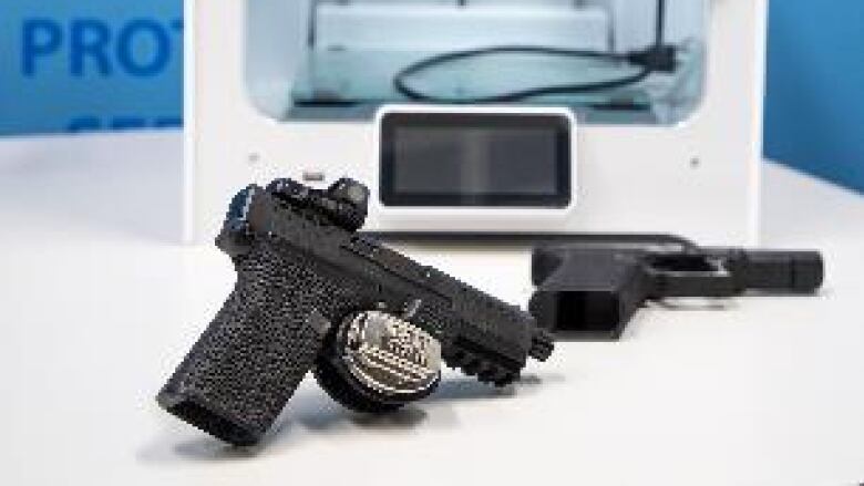 Two black handguns sit on a table, in front of a grey square-shaped printer.