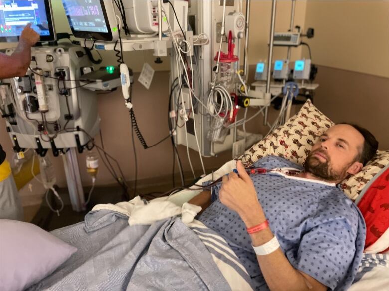 A man surrounded by hospital equipment lies in a hospital bed, looking at the camera.