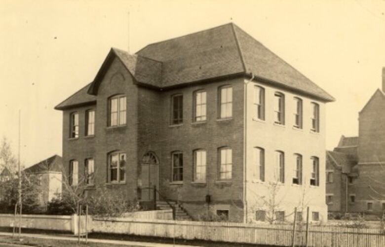 A very old black and white photo of a small square brick school building with a shingled roof.