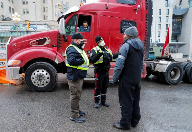 Two police officers talk to someone on a city street in winter. Someone sticks their head out of the window of a truck cab behind them to listen.