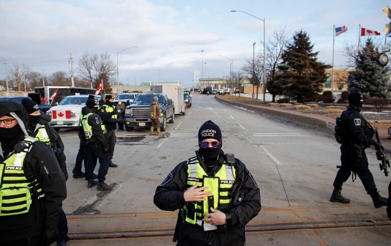 Police officers in bright neon vests stand in front of a row of pickup trucks with Canadian flags on them in front of a border crossing.