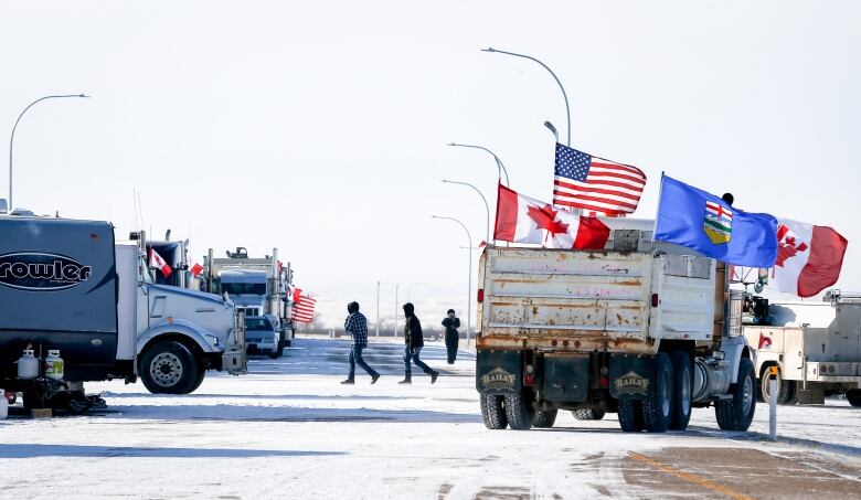 A number of large trucks and three people walking can be seen on a snowy road. A dump truck nearest the camera is flying large Canadian, Albertan and U.S. flags.