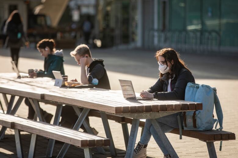 A male student sits at a picnic table at the University of British Columbia's Vancouver campus. He wears a facemask and has a laptop open on the table in front of him.