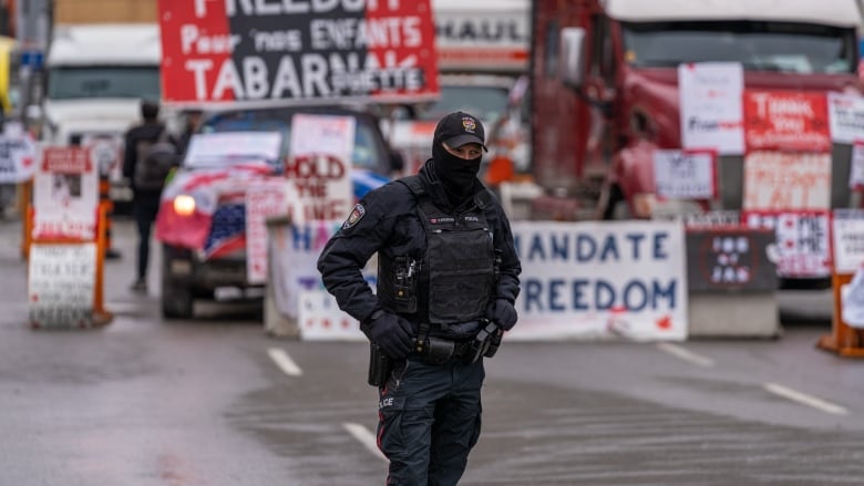 A uniformed officer with a balaclava stands on a street. In the rear are trucks and protest signs. 