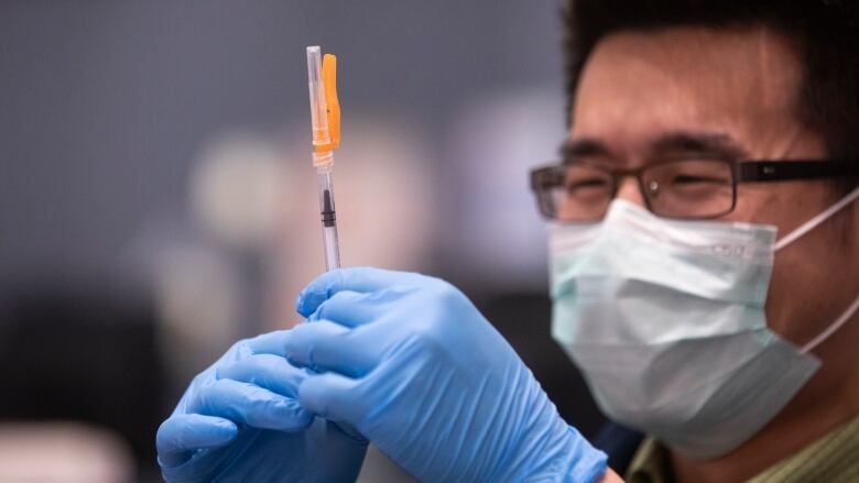 A man with a mask and blue gloves holds a syringe in his hand.