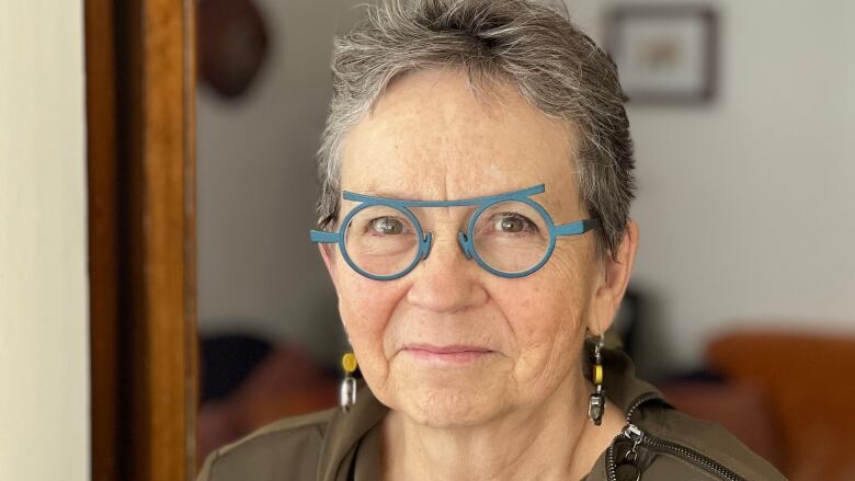 A woman with short hair and turquoise-coloured glasses smiles while looking at the camera.