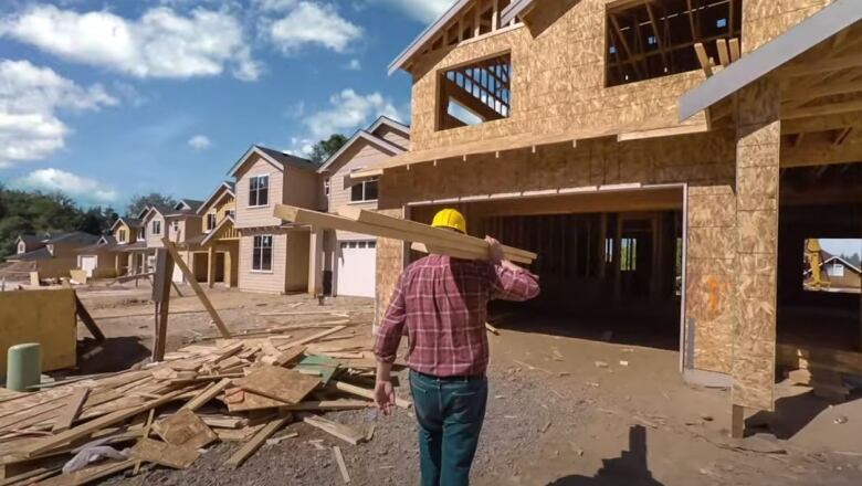 A construction worker viewed from behind wearing a red plaid shirt and carrying two-by-fours toward a partially completed house. 