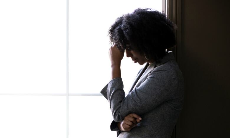 A Black woman stands in front of a window with a hand against her forehead, 