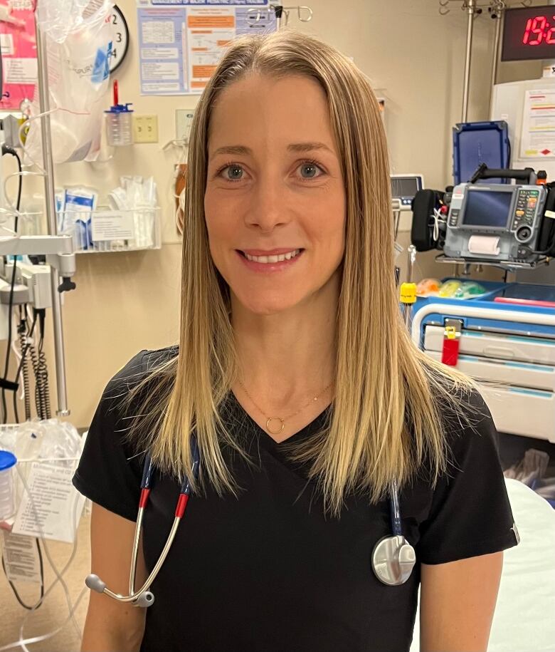 A doctor with long blonde hair stands in a hospital room with a stethoscope around her neck. 