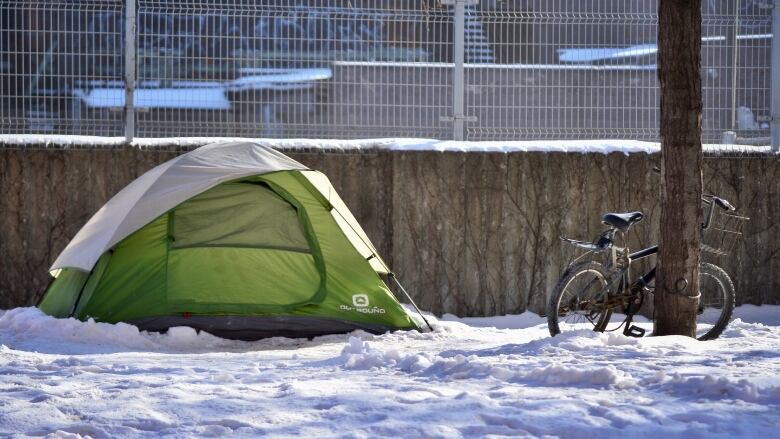 A green tent is set up in the snow with a bicycle nearby leaning against a tree.