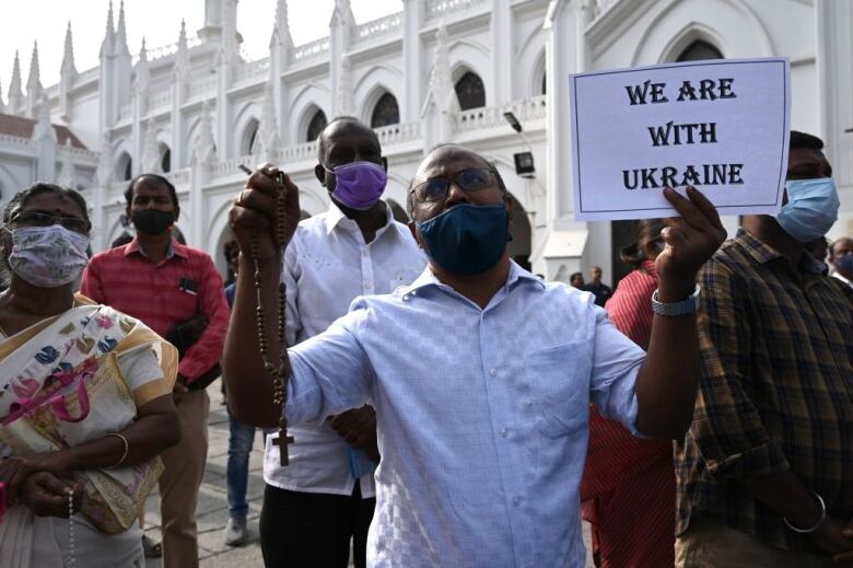 A group of people, some wearing masks, gather outside a large white church. A man in the foreground holds up a rosary and a sign that reads: We are with Ukraine.