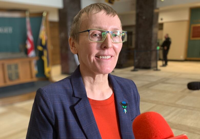 A person wearing a navy blazer, orange shirt and a brooch stands in the lobby of the Confederation Building.