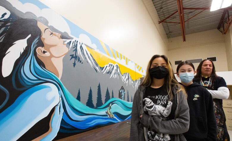 Three people pose in front of a mural that shows a flowing river, mountains and a sunny sky. The river appears as though it is flowing from a woman.