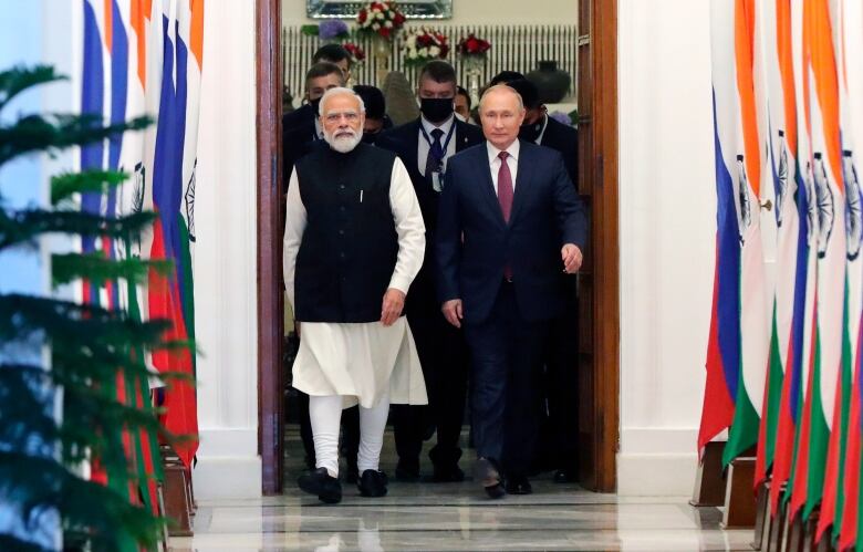 Two men walk side by side through a doorway, leading to a hall lined with flags.