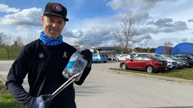 A man with a cap on his head and a blue scarf around his neck, wearing a blue coat, holds tongs and a small plastic bag of personal protective equipment he's picked up, while smiling for the camera.