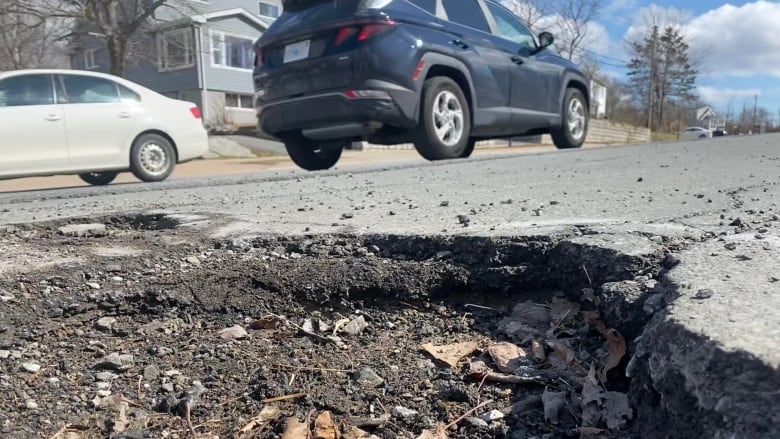 A large pothole filled with leaves and dirt on a road. Cars are behind it.