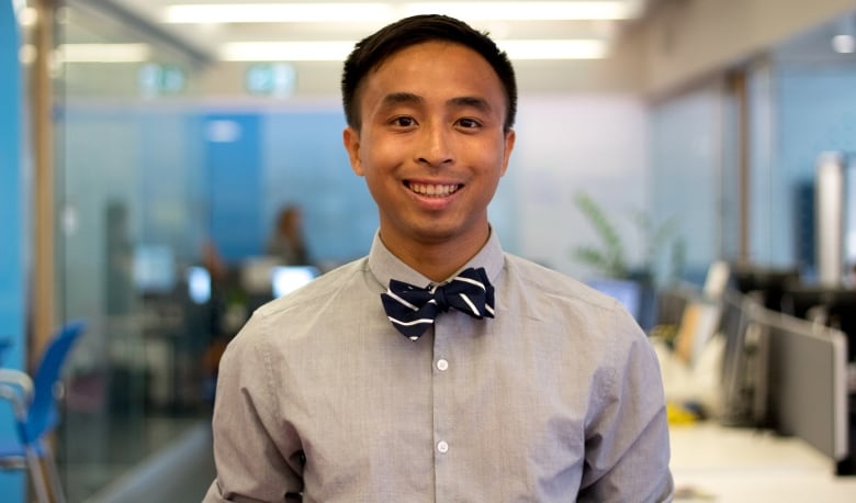 An Asian man with short hair wearing a grey shirt with a black striped bow tie stands in the middle of an open concept office.    