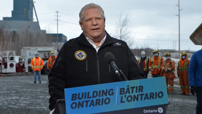 A man stands at a podium that says, Building Ontario, with workers in orange coveralls in the background.