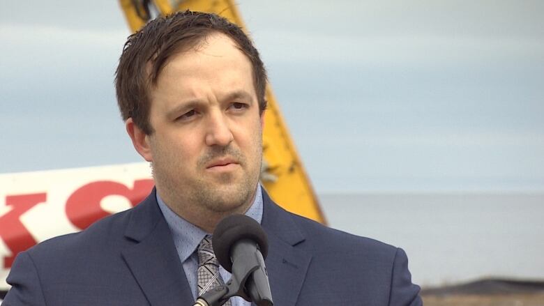 A man stands in front of a microphone outdoors. He is wearing a blue blazer with a blue shirt and a patterned tie.