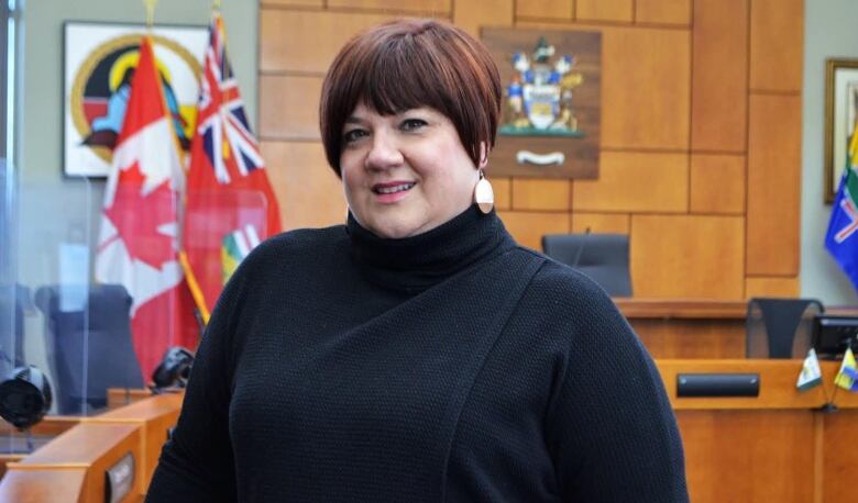 A person with short red hair wearing a dark-coloured sweater stands in a room, smiling.