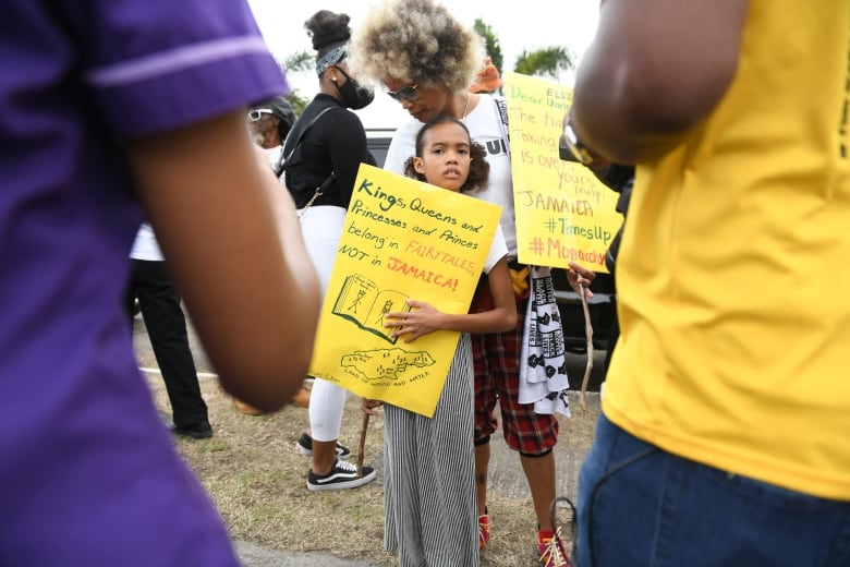 A young girl holding a yellow paper sign is embraced by a woman, as other demonstrators stand around them. The sign reads: Kings, Queens and Princesses and Princes belong in fairytales. Not in Jamaica.