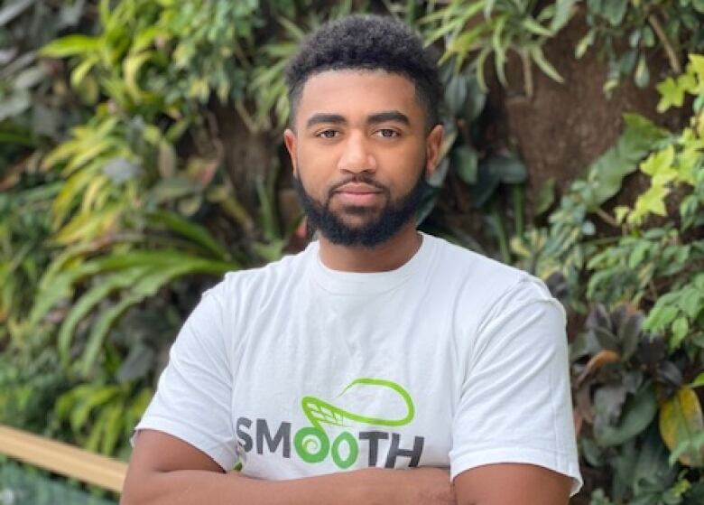 A Black man standing in front of a plant wall wearing a white t-shirt.