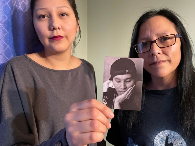 Two women hold up a photo of a young man in a ball cap. 