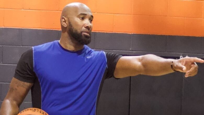 A bald Black man holding a basketball and wearing a black and blue shirt