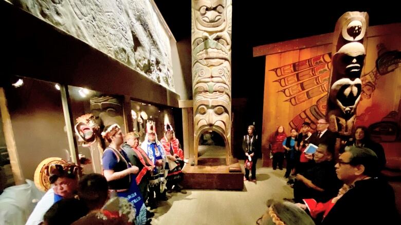 Members of the Nuxalk nation in ceremonial garb with other dignitaries present are arrayed around an intricately carved totem pole that is surrounded by native art in a museum.