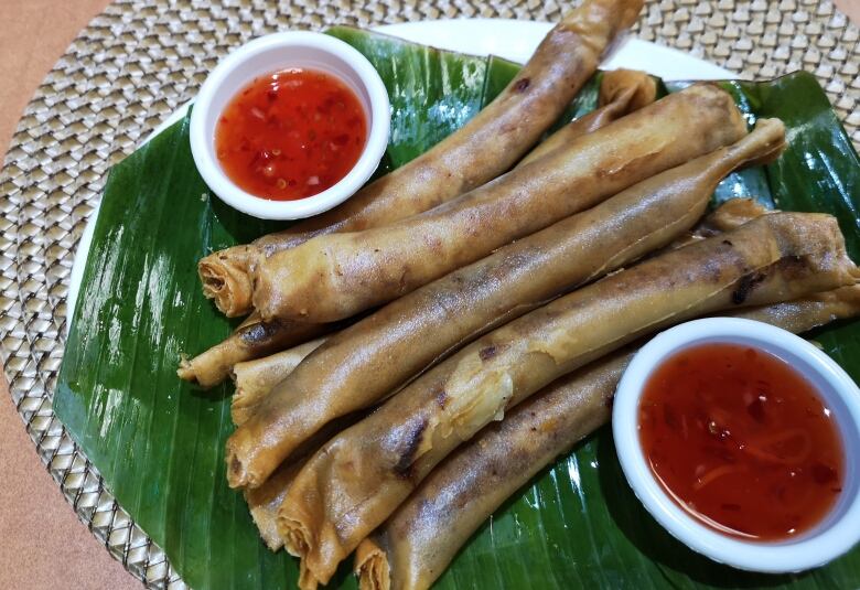 Thinly rolled spring rolls sit on a banana leaf, on top of a plate. Two bowls of dip. 