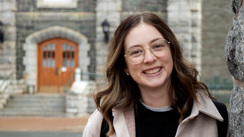 A smiling student with long brown hair and glasses poses in front of a building at Saint Mary's University in Halifax.