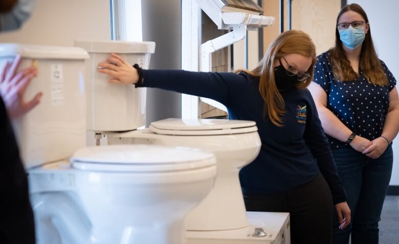 A student wearing a mask flushes one of two toilets on a dais in a large school room. 