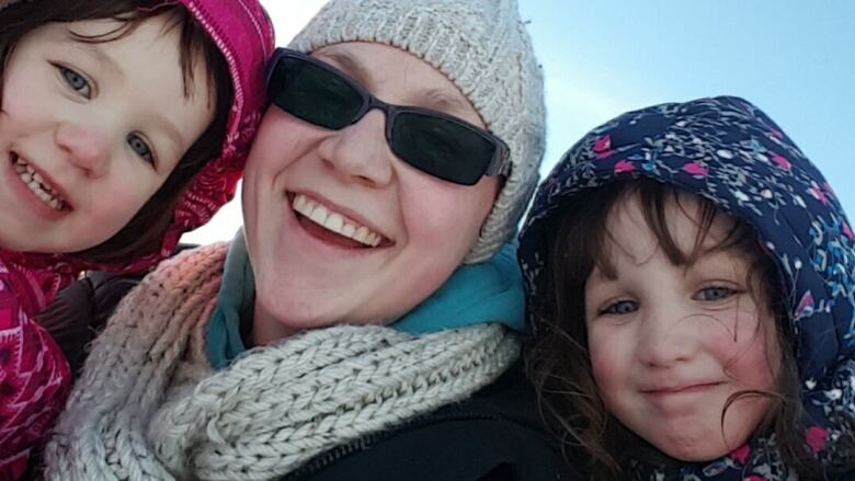 A closeup selfie shows a smiling woman wearing sunglasses, a tuque and a scarf standing outside on a sunny day between two young girls in winter coats with their hoods up.