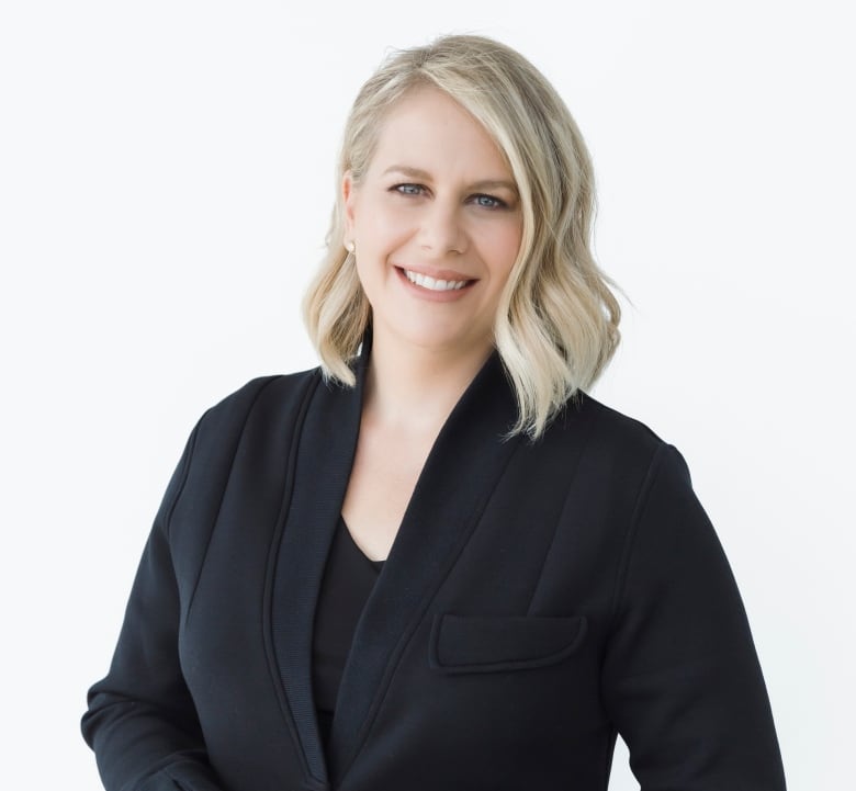 Host of Saskatoon Morning Leisha Grebinski stands in front of a white background wearing a black coat and a black shirt