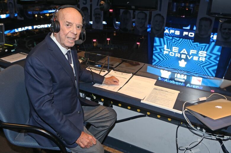 An older man wearing a suit sits in the broadcast booth at a hockey arena.