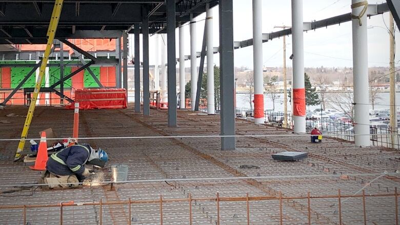 A large open area is shown inside a building under construction, with several large pillars holding up the ceiling and a man hunched over welding metal.