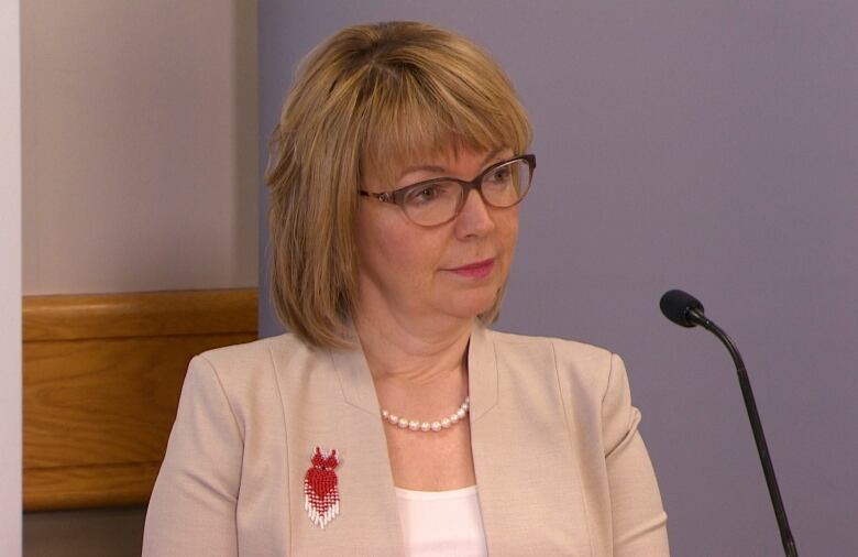 A woman with dyed blonde hair wearing a beige coloured jacket sitting at a microphone.