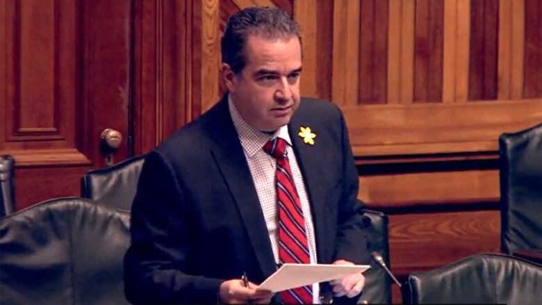 A man wearing a suit, holding papers while standing and speaking into a microphone.