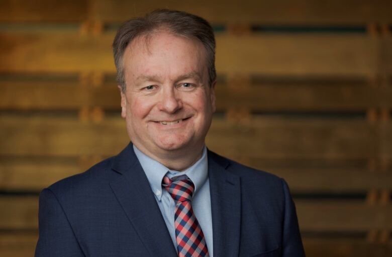 A man in his 40s or 50s with short brown hair and wearing a dark blue suit, light blue shirt and red and blue checked tied smiles at the camera for a head and shoulders professional portrait.