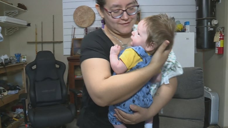 A woman wearing a black T-shirt and red glasses holds a toddler wearing a blue onesie with a print of Snoopy from the Charlie Brown series.