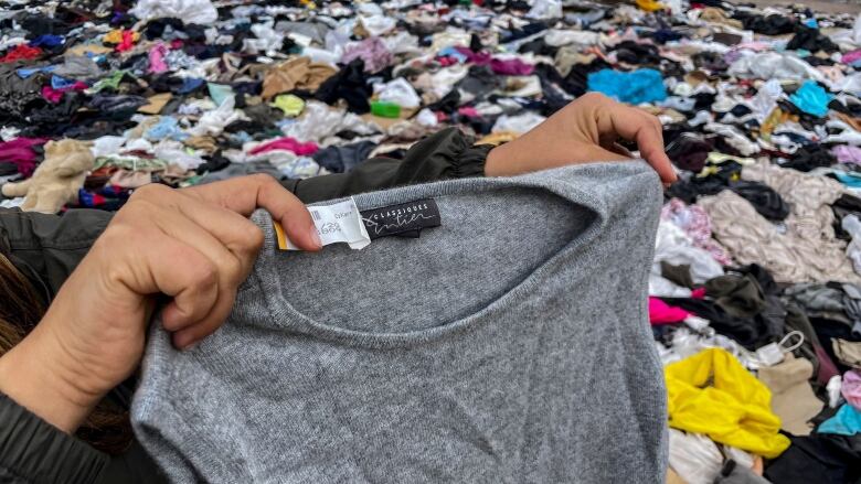 A pair of hands hold up a grey t-shirt against a backgroundstrewn with clothing.