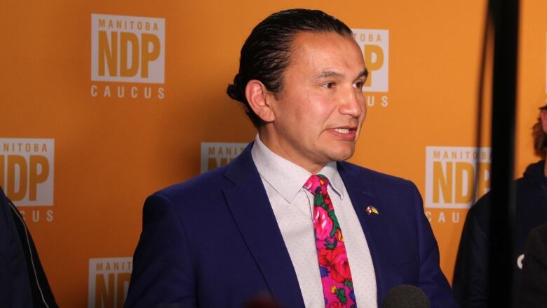 A man in a suit speaks at a podium, in front of a backdrop with the Manitoba NDP logo.