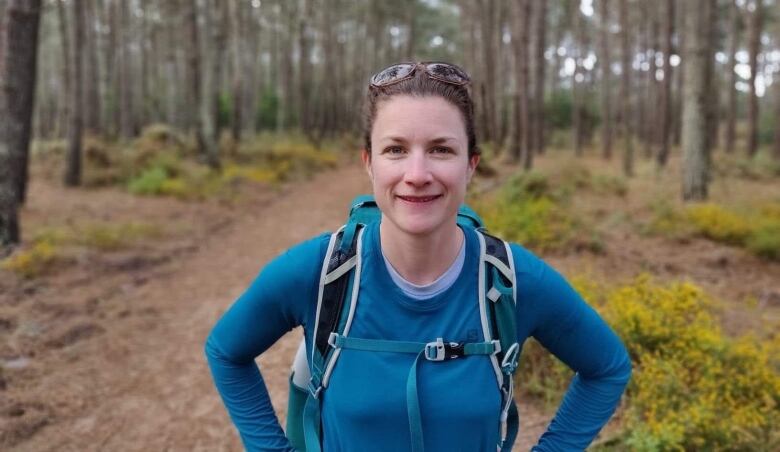 A woman with dark hair pulled back and dark sunglasses resting on her head stands on an outdoor wooded trail. She has a backpack on and is wearing a long-sleeve teal shirt.