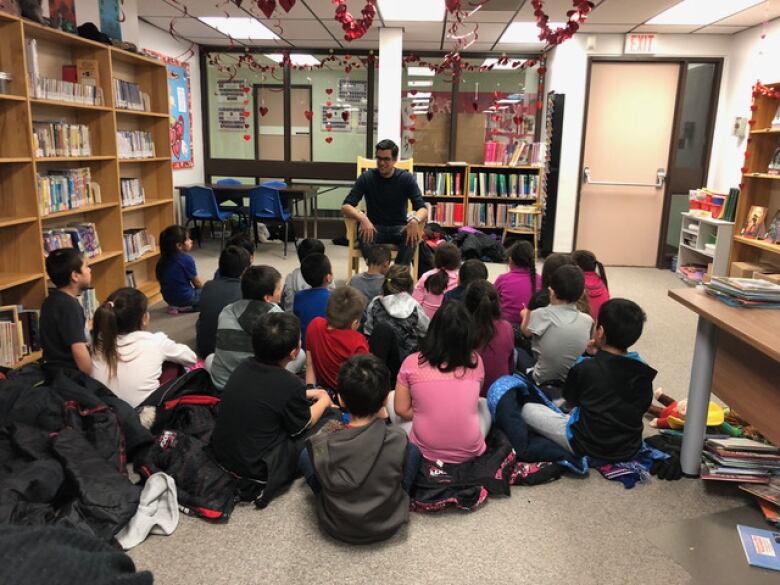 A man sits in a chair in a school library facing a group of schoolchildren, who sit cross-legged on the floor in front of him.  